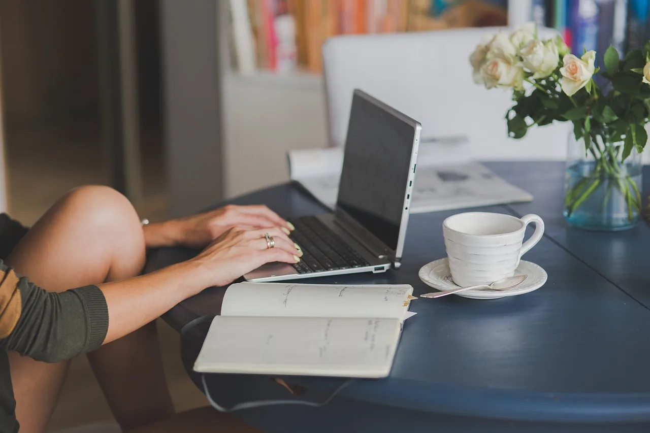 A woman typing on a laptop at a table with an open notebook, a cup of coffee and roses